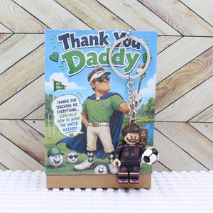 May include: A Father's Day gift set featuring a greeting card with the text "Thank You Daddy" and a golf-themed illustration. A soccer player Lego figure keychain is attached. The card includes the text "Thanks for teaching me everything... especially how to avoid the water hazard!"