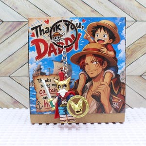 May include: A keychain featuring a cartoon figure with a sword and shield, hanging in front of a card with the words "Thank You, Daddy" and an illustration of two characters from the anime series One Piece.