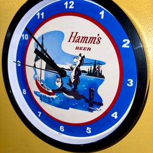 Puede incluir: Reloj de pared redondo con marco negro, esfera blanca y azul, y el logotipo de Hamm's Beer. La esfera del reloj presenta un oso de dibujos animados y una escena de lago. Las manecillas del reloj son negras.