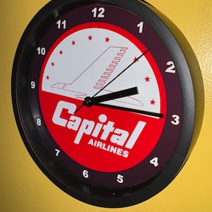 May include: Circular black wall clock with a red and white face. The clock face displays the words "Capital Airlines" and an aeroplane design. The clock has black hands and white numerals.