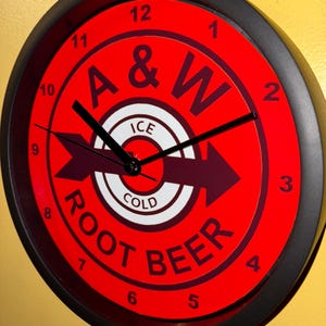 May include: A circular wall clock with a red face and a black frame. The clock face displays the A&W Root Beer logo, with the words "ROOT BEER" and "ICE COLD". Black hands show the time.