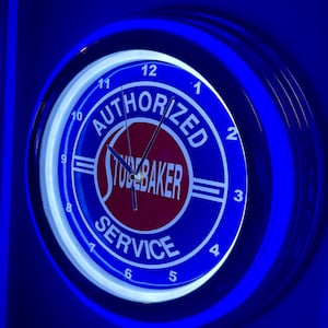 Puede incluir: Un reloj de neón redondo, azul y rojo, con el texto "AUTHORIZED STUDEBAKER SERVICE". La esfera del reloj tiene números y manecillas blancas, y está rodeada por una luz de neón azul. El reloj está montado en una pared azul.