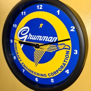 May include: Circular wall clock with a black surround. The clock face is blue and yellow, displaying the text "Grumman Aircraft Engineering Corporation". White numerals indicate the hours.