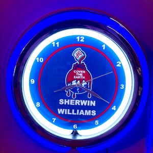May include: A round clock with a blue and white neon light border. The clock face is blue with the words "SHERWIN WILLIAMS" and a logo. The clock hands are black. The clock is illuminated with a blue light.