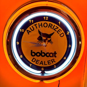 Puede incluir: Un reloj de neón redondo con una esfera amarilla y un borde negro y amarillo. La esfera del reloj muestra las palabras "AUTHORIZED" y "bobcat DEALER" con un logotipo de lince. El reloj está enmarcado en naranja.