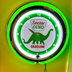 Puede incluir: Un reloj de pared redondo e iluminado con un borde de neón verde. La esfera del reloj presenta el logotipo de Sinclair Oil con un dinosaurio verde y la palabra "GASOLINE". El reloj tiene manecillas negras y un fondo blanco con números negros.