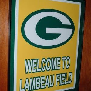 Puede incluir: Un letrero enmarcado con fondo amarillo y un logotipo ovalado verde y blanco. El letrero dice "WELCOME TO LAMBEAU FIELD" en letras verdes y blancas. El marco es negro.