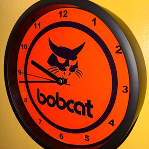 Puede incluir: Un reloj de pared redondo con marco negro y esfera naranja. La esfera del reloj presenta la palabra "bobcat" en blanco y un logotipo de lince negro. Las manecillas negras indican la hora.