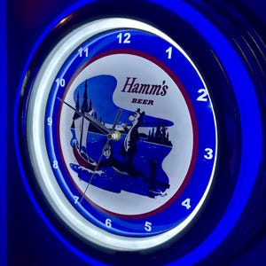 Puede incluir: Un reloj redondo e iluminado de Hamm's Beer con un diseño azul y blanco. La esfera del reloj presenta un oso en un barco en un lago. El reloj tiene un borde de neón azul y muestra los números del 1 al 12.