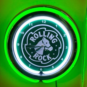 May include: A round, green neon clock with the words "ROLLING ROCK" and a horse head logo. The clock face is white with black numbers and hands. The clock is surrounded by a green neon light.