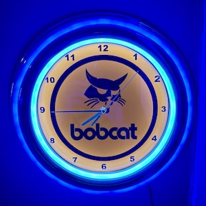 Puede incluir: Un reloj de pared redondo con una esfera amarilla y un borde de luz de neón azul. La esfera del reloj presenta la palabra "bobcat" debajo de una silueta negra de la cabeza de un gato montés. Las manecillas del reloj son azules.