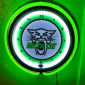 May include: A round, neon green Arctic Cat clock. The clock face is white with black numbers and the Arctic Cat logo in the center. The words "ARCTIC CAT" are in green. The clock is surrounded by a bright green neon light.