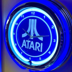 May include: A round, blue neon Atari clock with a silver-colored rim. The clock face is dark blue with the Atari logo and white numerals. The clock has a bright blue neon light around the face and the outer rim. The hands are silver.