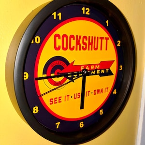 May include: Round wall clock with a black frame and a yellow face. The clock face features the word "COCKSHUTT" in red, with the words "FARM MENT" and "SEE IT US IT OWN IT". The clock has black hands and yellow numbers.