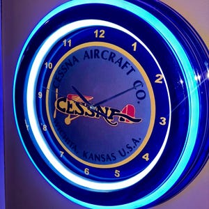 May include: A circular blue neon clock displaying the text "Cessna Aircraft Co." and an airplane graphic. The clock face is blue with yellow numerals and hands. The outer rim is illuminated with a bright blue neon light.