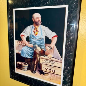 May include: Framed vintage advertisement for J.B. Lewis, Shoe Maker, Boston. The image features a man in a blue apron with the text "WEAR RESISTERS" and a wooden box with the text "WORKING FOR YOU". The frame is black with a speckled design.