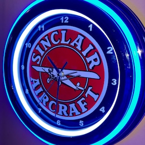 May include: A circular wall clock with a blue neon surround and a white neon inner ring. The clock face displays the words "Sinclair Aircraft" around a red circle with a white aeroplane graphic. Black clock hands show the time.