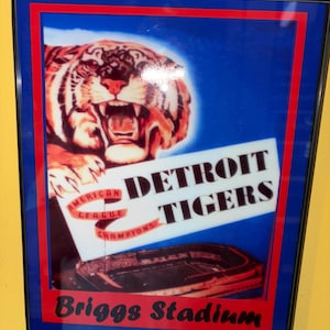 Puede incluir: Póster vintage enmarcado de los Detroit Tigers. El póster presenta un tigre rugiente, las palabras "Detroit Tigers" y "Briggs Stadium". El fondo es azul con detalles en rojo y blanco. El póster está enmarcado en negro.