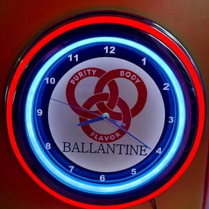 Puede incluir: Un reloj de pared redondo con un borde de luz de neón rojo y azul. La esfera del reloj es blanca con la palabra "BALLANTINE" y las palabras "Purity, Body, Flavor". Las manecillas indican las 2:10.