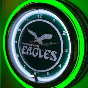 Könnte beinhalten: Eine runde Wanduhr mit grün-weißem Zifferblatt mit dem Logo der Philadelphia Eagles. Die Uhr hat einen neongrünen Rand und ein weißes Neonlicht um das Zifferblatt. Die Zeiger der Uhr sind schwarz.