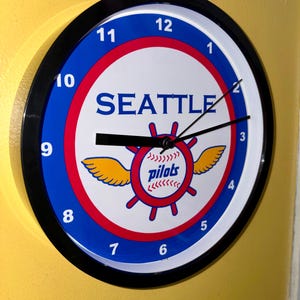 May include: Circular wall clock with a black surround. The white face displays blue numerals and the word "SEATTLE". The centre has a red and white design with the word "pilots" and yellow wings.