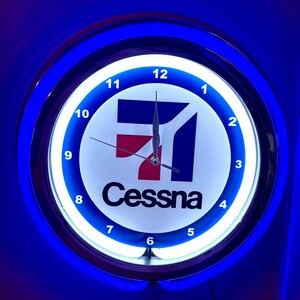May include: A round, illuminated wall clock with a blue and white design. The clock face features the Cessna logo in red, white, and blue, with black hands and numerals. The clock is surrounded by a blue neon light.