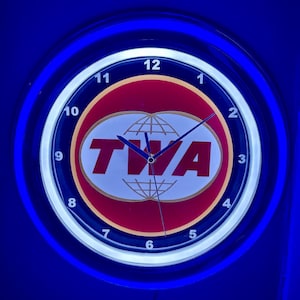 May include: A circular wall clock with a blue neon light surround. The clock face displays the TWA logo in red and white, with a globe graphic. Black numerals mark the hours. The clock hands are black and indicate 1:50.
