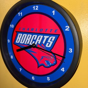 Puede incluir: Un reloj de pared redondo con marco negro. La esfera tiene un centro rojo con el texto "CHARLOTTE BOBCATS" y un anillo exterior azul con números blancos. Las manecillas indican aproximadamente las 8:15.
