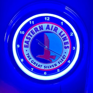 May include: A circular, illuminated clock with a blue surround and a white inner ring. The clock face displays the text "EASTERN AIR LINES" and "THE GREAT SILVER FLEET" around a central emblem of a red bird in flight. The clock hands indicate 10:10.