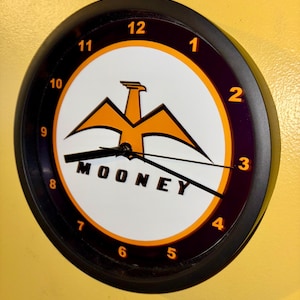 May include: A circular wall clock with a black surround and a white face. The clock displays an orange eagle logo above the black word "MOONEY". The numerals are orange, and the hands are black.