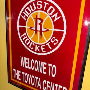 Op de afbeelding: Rood bord met het logo van de Houston Rockets, een basketbal met een gestileerde "R". De tekst "Welcome to the Toyota Center" is wit. Het heeft een zwarte lijst en gele en oranje accenten.