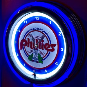 Peut inclure: Une horloge ronde et lumineuse avec une bordure néon bleue. Le cadran affiche le logo des Phillies de Philadelphie en rouge et blanc, avec les mots "PHILADELPHIA" et "Phillies". Les aiguilles indiquent l'heure.