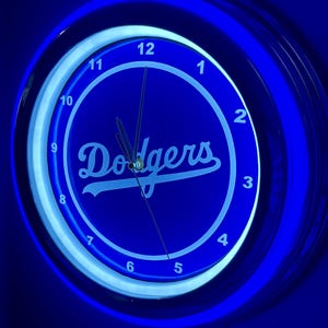 May include: A blue neon clock with the word "Dodgers" in white script. The clock face has white numerals and hands, and is surrounded by a blue neon light. The clock is a sports-themed item.