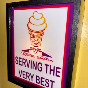 Puede incluir: Letrero luminoso de Mister Softee con marco negro. El letrero presenta un cono de helado de dibujos animados con una cara sonriente y el texto "SERVING THE VERY BEST" en morado.