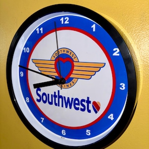 May include: Circular wall clock with a black surround, displaying the Southwest Airlines logo. The clock face is white with blue and red detailing, showing the time with black hands and numerals.