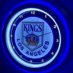 May include: A round, illuminated wall clock with a blue neon border. The clock face features the words "KINGS LOS ANGELES" around a team logo with a crown. The clock hands are black.