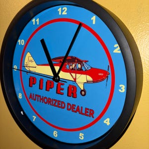 May include: Round wall clock with a black frame, featuring a blue face with white numerals and black hands. The clock face displays a red and yellow airplane illustration with the text "PIPER AUTHORIZED DEALER".