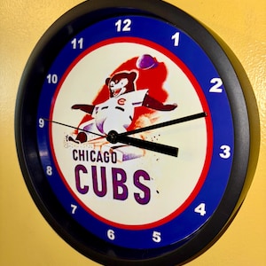 May include: Round wall clock with a black frame, featuring the Chicago Cubs logo. The clock face has a white background with a red and blue design, and the Cubs bear mascot. The clock hands are black.