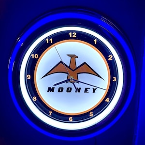 May include: A circular wall clock with a blue neon border. The clock face is white with black numerals and hands. The centre features an orange eagle design above the word "MOONEY".