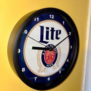 May include: Round wall clock with a black frame and a white face featuring the word "Lite" in navy blue. The clock face also includes the words "AF PILSENER BEER" and a red and gold emblem. The clock is mounted on a yellow wall.