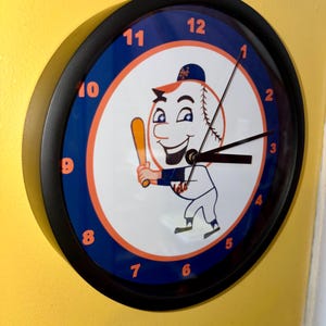 May include: A round wall clock with a black frame. The clock face features a baseball with a cartoon character holding a bat. The numbers are in blue, and the background is white and orange. The clock is mounted on a yellow wall.