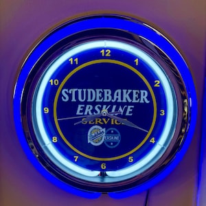 Puede incluir: Un reloj de pared redondo e iluminado con un bisel azul y cromado. La esfera del reloj es azul oscuro con las palabras "STUDEBAKER ERSKINE SERVICE" en blanco y amarillo. El reloj tiene un anillo exterior azul neón y un anillo interior blanco.