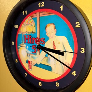 Puede incluir: Reloj de pared redondo negro con un anuncio vintage de Hires 5¢. La esfera del reloj tiene un borde rojo y azul, con un hombre sonriente con traje blanco. Las manecillas del reloj son negras y los números blancos.