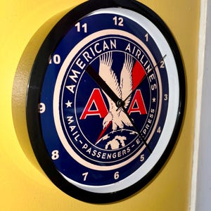 May include: A round wall clock with a black frame. The clock face features the American Airlines logo in blue, red, and white, with the words "American Airlines Mail-Passengers-Express" around the edge. The clock hands are black.