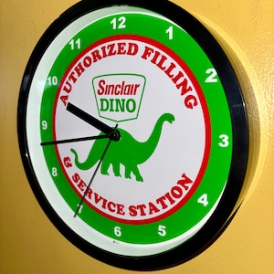Puede incluir: Reloj de pared redondo con marco negro. La esfera blanca presenta detalles verdes y rojos, mostrando el logotipo de Sinclair Dino y el texto "AUTHORIZED FILLING & SERVICE STATION". Las manecillas negras indican la hora.