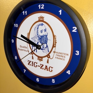May include: A round wall clock with a black frame and a white face. The clock face features a blue and brown design with the text "ZIG-ZAG" and an illustration of a man with a beard. The clock hands point to 9:50.
