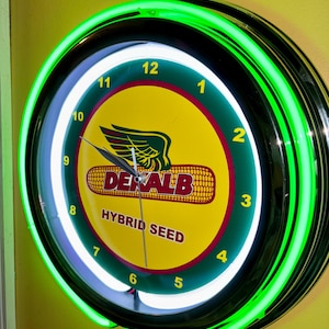May include: A round, neon clock with a black frame and a bright green neon outer ring. The clock face is yellow with the words "DEKALB HYBRID SEED" and a corn graphic. Numbers 1-12 are visible.