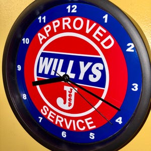 May include: Circular wall clock with a black surround. The clock face has a red and blue design with the words "APPROVED WILLYS SERVICE" in white. The clock hands are black, and the numbers are white.