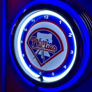 May include: A neon wall clock with a blue and white Phillies logo. The clock has a blue neon border and a white neon inner ring. The clock face displays the numbers 1 through 12. The hands of the clock are black.
