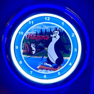 Puede incluir: Un reloj redondo e iluminado con un anillo exterior azul y un anillo interior blanco. La esfera del reloj presenta un oso de dibujos animados y la palabra "Hamms" en rojo. El fondo representa un lago y una escena de bosque.